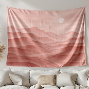Peut inclure: Une grande tapisserie rectangulaire avec un paysage de style aquarelle dans des tons corail et pêche est suspendue au-dessus d'un canapé blanc. L'œuvre représente des collines ondulantes superposées avec un soleil blanc. La pièce est décorée dans des tons neutres.