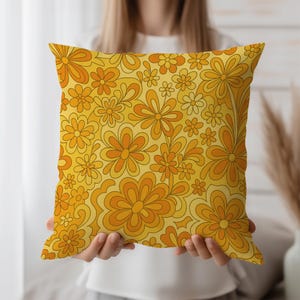 May include: A square throw pillow with a retro floral pattern. The design features orange and yellow flowers with a groovy, 1970s aesthetic. The pillow is held up by a person.