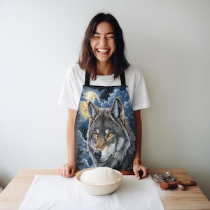 Puede incluir: Un delantal negro con un diseño de lobo aullando a la luna. El lobo es gris y marrón con ojos amarillos. La luna es amarilla y el fondo es azul oscuro con nubes blancas.