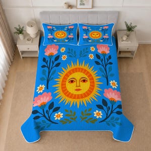 May include: A blue quilt and two matching pillow shams featuring a large central yellow and orange sun with a face, surrounded by pink flowers, green leaves, and small white blossoms. The bedding set has a white border trim on all edges.