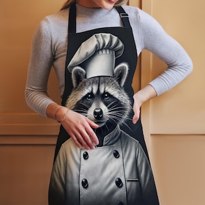 Könnte beinhalten: Schwarze Schürze mit einer weißen Kochmütze und einem Waschbären, der eine Kochuniform trägt. Der Waschbär hat eine schwarze Nase und schwarze Augen.