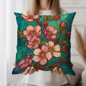 Puede incluir: Un cojín cuadrado decorativo con un diseño floral estilo vidriera. El fondo turquesa presenta flores de cerezo rosas y ramas marrones. Las flores tienen detalles dorados. Un accesorio decorativo para el hogar.