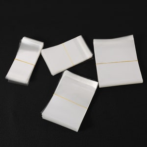 May include: Multiple stacks of clear cellophane bags with white backing, secured with yellow elastic bands. The bags are arranged on a black surface, with some stacks overlapping each other.