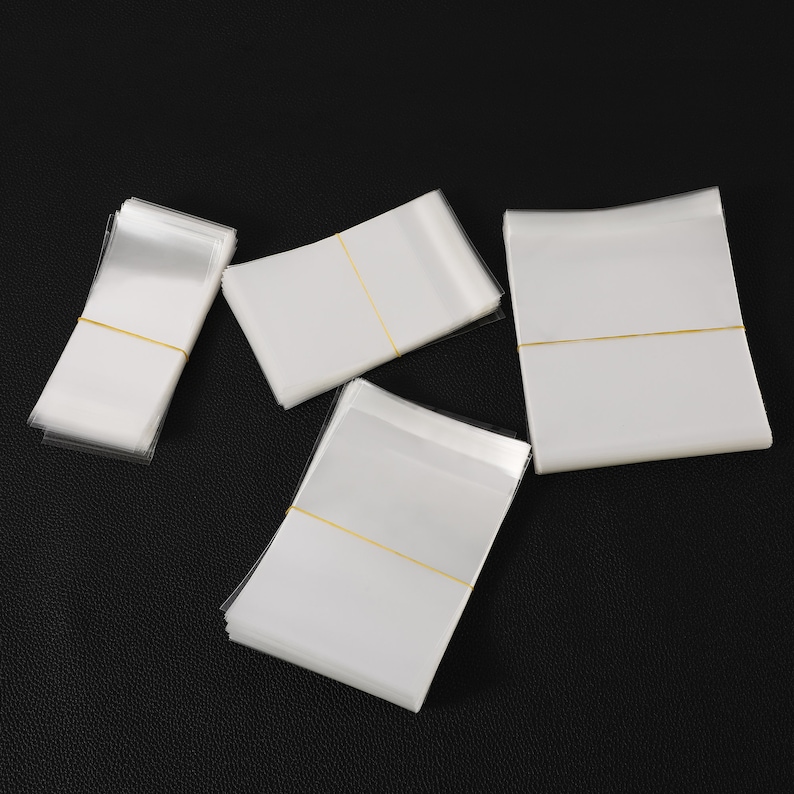 May include: Multiple stacks of clear cellophane bags with white backing, secured with yellow elastic bands. The bags are arranged on a black surface, with some stacks overlapping each other.