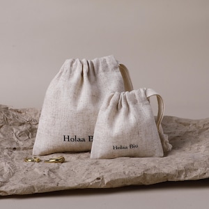 以下が含まれることがあります： 「Holaa B」と「Holaa Biu」の文字が黒でプリントされた、ベージュ色の巾着袋が2つ。2つの金の指輪が袋の前にあります。袋は、質感のある薄茶色の表面に置かれています。