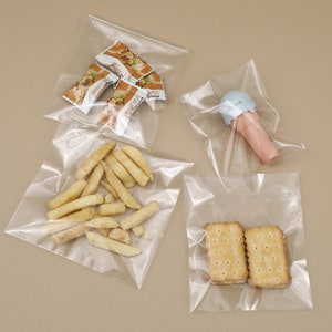 May include: Four clear plastic bags, each containing different snacks. One bag holds wrapped sweets, another has french fries, a third contains an ice cream toy, and the last one has two biscuits.