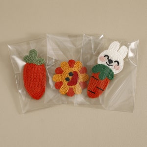 May include: Three crocheted appliques in clear plastic bags. The appliques include a red strawberry with a green top, a yellow and orange sunflower, and a white bunny with a carrot. The bunny has pink cheeks and a smiling face.