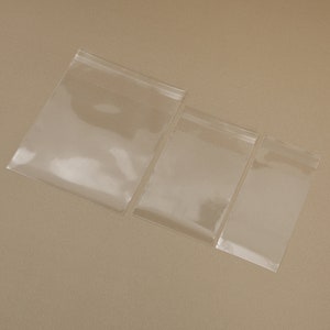 May include: Three clear, transparent plastic zip-top bags of varying sizes are arranged diagonally on a neutral background. The bags are empty and sealed, showcasing their simple design and functionality. The bags are ideal for storing small items.