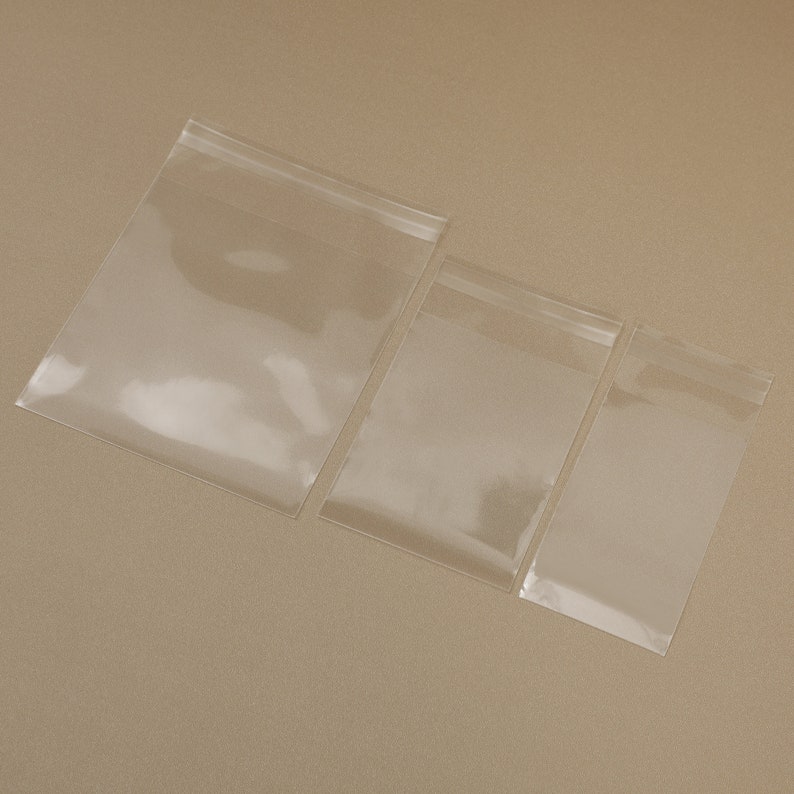 May include: Three clear, transparent plastic zip-top bags of varying sizes are arranged diagonally on a neutral background. The bags are empty and sealed, showcasing their simple design and functionality. The bags are ideal for storing small items.
