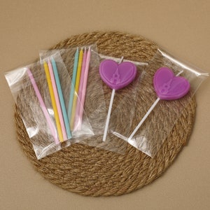May include: A selection of candles and cake toppers in clear plastic packaging. The candles are in pastel shades of pink, yellow, and blue. The cake toppers are heart-shaped, purple, with a castle design. They are displayed on a woven mat.