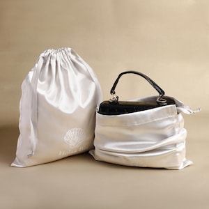 Pode incluir: Duas sacolas brancas de cetim com cordão, uma contendo uma bolsa preta. As sacolas têm um desenho de concha e o texto "Holaa Bii". A bolsa tem uma alça preta e ferragens douradas.