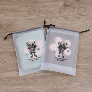 May include: Two white drawstring bags with a black and white cartoon character design. The character is a creature with a large head and a small body. The text on the bag reads "YOU'RE SO PRETTY!" and "YOU'RE SO PRETTY!"