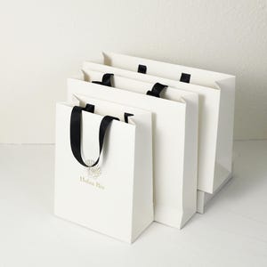 Könnte beinhalten: Vier weiße Geschenktüten mit schwarzen Schleifenhenkeln. Die Taschen sind rechteckig und in einer Reihe angeordnet. Auf der vorderen Tasche steht der Text "Holaa Biu" in Gold gedruckt, zusammen mit einer goldenen Grafik.