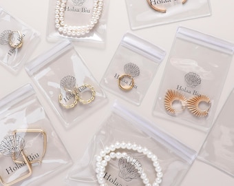 Puede incluir: Una colección de joyas que incluye collares de perlas, pendientes de aro dorados y anillos, empaquetados individualmente en bolsas de plástico transparente. Cada bolsa lleva el logotipo de Holaa Biu con un gráfico de concha en negro.