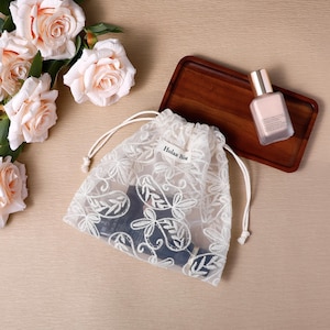 May include: A white sheer drawstring pouch featuring an intricate floral embroidery pattern and a small fabric tag labeled "Holaa Biu". The pouch is positioned next to a rectangular wooden tray holding a glass bottle with a metallic gold cap.