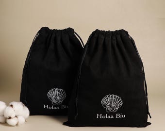 May include: Two black drawstring bags with white seashell logos and the text "Holaa Biu". The bags are made of a soft material and have a drawstring closure. A few cotton buds are visible on the left side of the image.