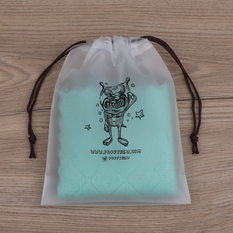 May include: A clear plastic drawstring bag with a teal blue fabric lining. The bag features a black and white illustration of a cartoon monster with stars and the text "www.puppysew.com @puppysew".