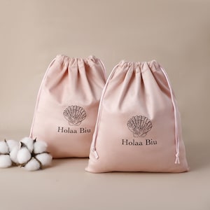 Pode incluir: Duas sacolas rosa blush com cordão com um desenho de concha e o texto "Holaa Biu". As sacolas são feitas de um material macio e têm um fecho de cordão. Um grupo de botões de algodão está em primeiro plano.