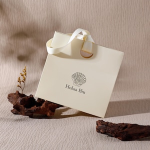May include: A square, cream-colored gift bag with a white ribbon handle and a semi-circular wooden accent. The front features a black line-art illustration of a seashell centered above the text "Holaa Biu" in a clean, black serif font.