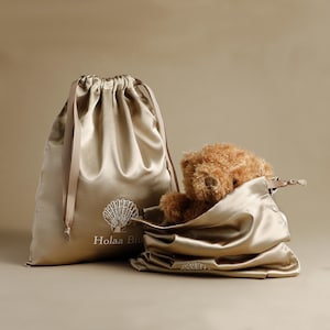 Puede incluir: Dos bolsas de satén con cordón en color champán, una grande y otra pequeña, con un osito de peluche asomando por la bolsa más pequeña. La bolsa grande tiene un diseño de concha y el texto "Holaa Bitu".