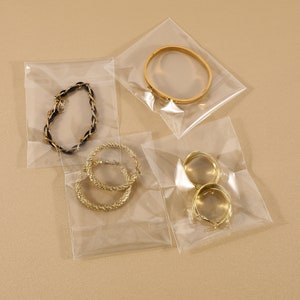May include: Four pieces of jewellery are displayed in clear plastic bags. The jewellery includes a gold bracelet, gold hoop earrings, and a black and gold braided bracelet. The jewellery is on a neutral background.