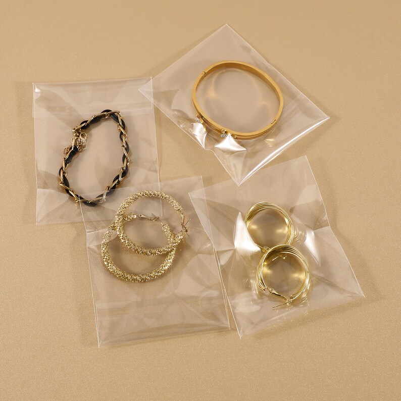 May include: Four pieces of jewellery are displayed in clear plastic bags. The jewellery includes a gold bracelet, gold hoop earrings, and a black and gold braided bracelet. The jewellery is on a neutral background.