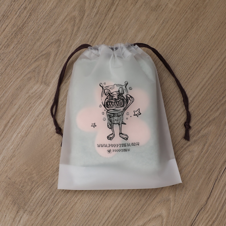 May include: A white drawstring bag with a black and white illustration of a cartoon character wearing a space helmet. The character is holding a star and has a big smile. The text "www.poppysew.com" and "@poppysew" is printed below the illustration.