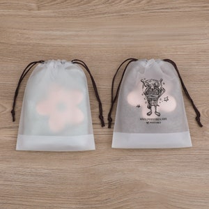 May include: Two white drawstring bags with brown drawstrings. One bag has a pink flower design, the other has a black and white illustration of a cartoon monster with the text "SMALL JOYFUL THINGS" and "TO PURSUE".