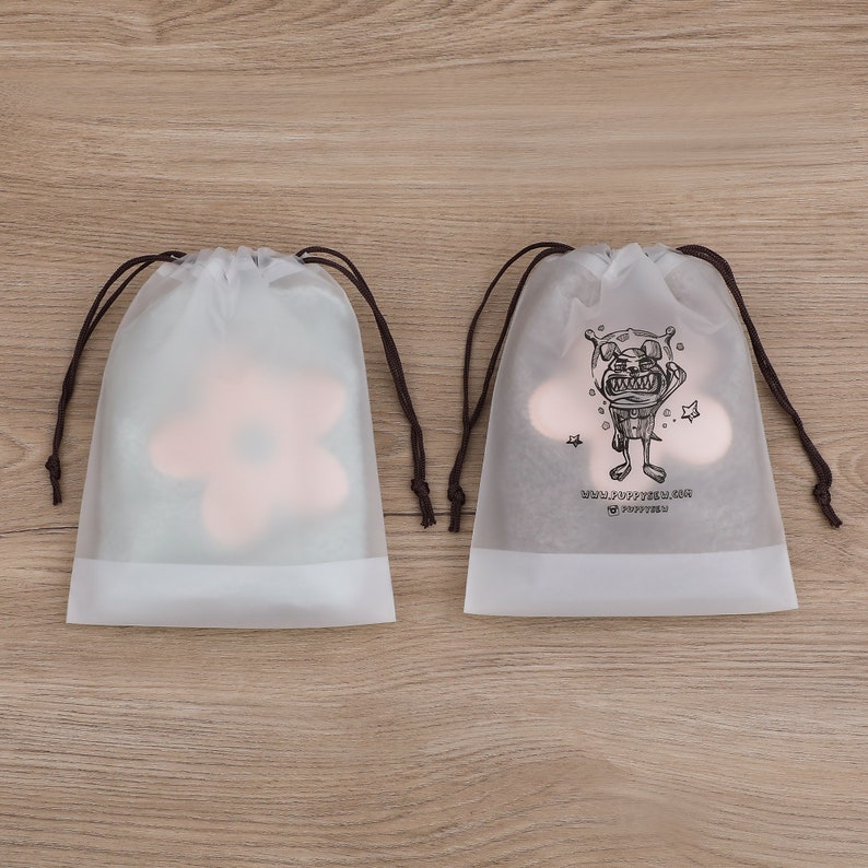 May include: Two white drawstring bags with brown drawstrings. One bag has a pink flower design, the other has a black and white illustration of a cartoon monster with the text "SMALL JOYFUL THINGS" and "TO PURSUE".