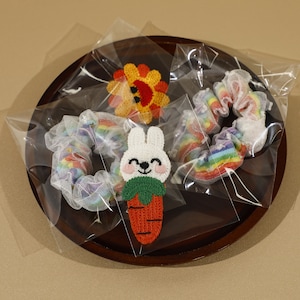 May include: A selection of handmade hair accessories, including rainbow scrunchies and a bunny-shaped patch with a carrot. The items are packaged in clear plastic and presented on a wooden tray. These accessories are likely for children.