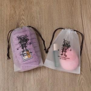 May include: Two clear plastic drawstring bags with black drawstrings. Each bag has a cartoon illustration of a monster with the text "YOU'RE SO PRETTY, DEAR" and "YOU'RE SO PRETTY". One bag contains a purple knitted hat and the other contains a pink makeup sponge.