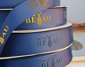 May include: A stack of navy blue grosgrain ribbon rolls featuring the text MAISON BEAU and a bee icon. The top two rolls display the logo in metallic gold foil, while the bottom two rolls feature the same design in a dark, matte finish.
