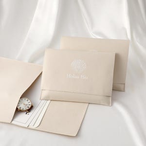 May include: Beige pouches with a seashell design and the text "Holaa Biu". One pouch is open, revealing a watch with a brown leather strap and some cards. The pouches are on a white, silky surface.