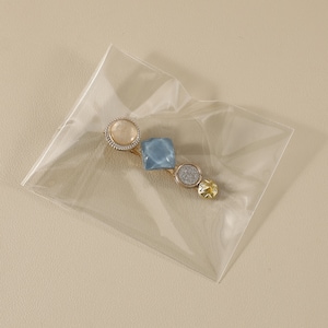 May include: A gold-coloured hair barrette with decorative elements. It features a round, light-coloured gem, a square blue gem, a round silver glitter gem, and a star-shaped gold gem. The barrette is in a clear plastic package.