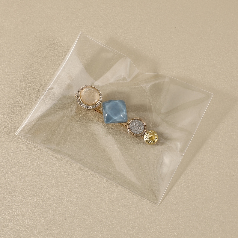 May include: A gold-coloured hair barrette with decorative elements. It features a round, light-coloured gem, a square blue gem, a round silver glitter gem, and a star-shaped gold gem. The barrette is in a clear plastic package.