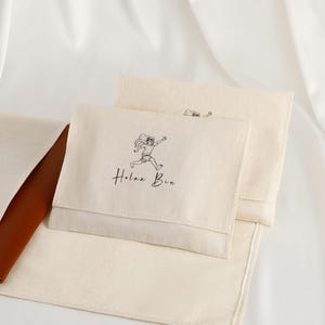 Könnte beinhalten: Off-white Canvas-Beutel mit einer Illustration einer laufenden Figur und dem Text "Holaa Bin" in Schwarz. Die Beutel sind rechteckig mit einem Klappenverschluss. Ein brauner Lederartikel ist teilweise sichtbar.