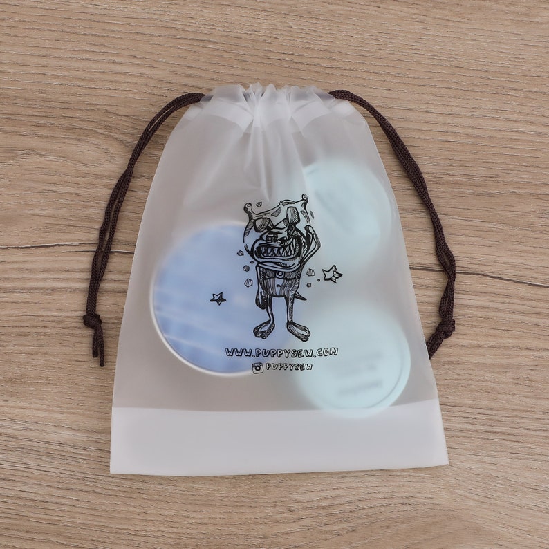 May include: A clear plastic drawstring bag with a black and white cartoon monster graphic. The bag has a brown drawstring and the text "www.puppysew.com @puppysew" printed on the front.