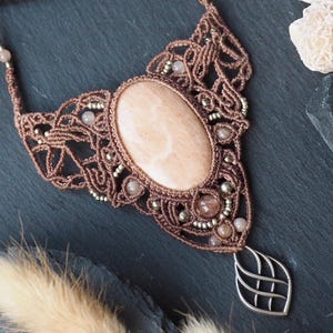 May include: A handcrafted macrame necklace with a large, oval, peach-colored stone centerpiece. The intricate brown macrame design is accented with small beads and a silver leaf-shaped pendant. The necklace is displayed on a dark surface.