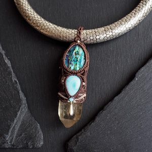 Könnte beinhalten: Eine handgefertigte Halskette mit einem Anhänger im braunen Makramee-Design. Der Anhänger enthält eine blau-grüne Abalone-Schale, einen hellblauen Stein und einen klaren Quarz-Kristall. Die Halskette hat eine silberfarbene, strukturierte Kette.