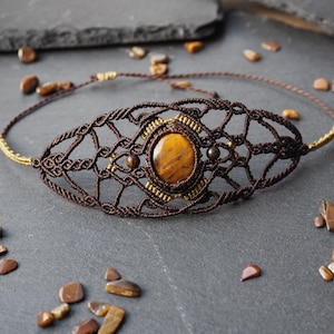 May include: A macrame choker necklace with a central tiger's eye stone. The necklace has a brown woven design with gold accents and small beads. The tiger's eye stone is oval-shaped and has a warm, golden-brown colour. The necklace is suitable for a neck circumference of approximately 35 cm.