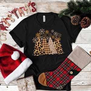 Pu&ograve; includere: Maglietta nera con un design di tre alberi di Natale decorati con stampa leopardata, a quadri e glitter. Gli alberi sono circondati da fiocchi di neve.