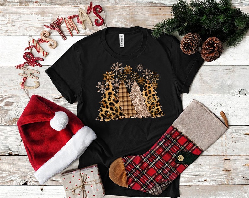 Pu&ograve; includere: Maglietta nera con un design di tre alberi di Natale decorati con stampa leopardata, a quadri e glitter. Gli alberi sono circondati da fiocchi di neve.