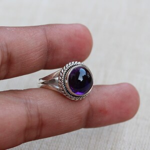 May include: A silver ring with a large, round, deep purple gemstone. The gemstone is set in a circular bezel with a braided rope design around it. The ring band splits into two parts on each side of the gemstone.