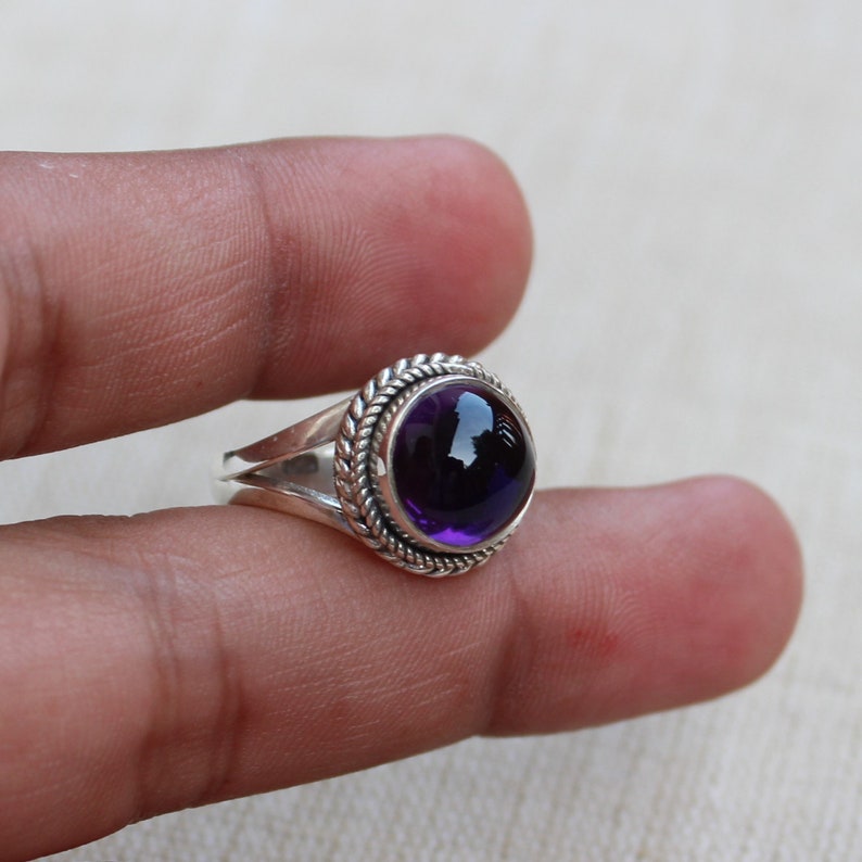 May include: A silver ring with a large, round, deep purple gemstone. The gemstone is set in a circular bezel with a braided rope design around it. The ring band splits into two parts on each side of the gemstone.
