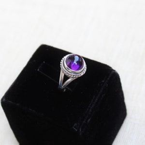 May include: A silver ring with a large, round, deep purple gemstone. The gemstone is set in a decorative bezel with a twisted rope design. The ring is displayed on a black velvet surface.