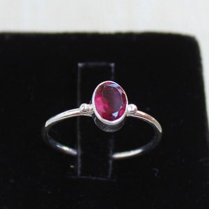Pode incluir: Um anel de prata com uma pedra preciosa ovalada de cor vermelha escura. O anel tem uma banda simples com pequenos detalhes decorativos em ambos os lados da pedra preciosa. A pedra é definida em uma configuração de moldura.