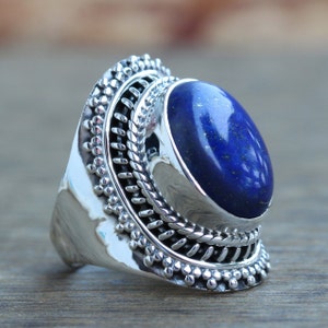 Peut inclure: Bague en argent ornée d'une grande pierre ovale de lapis-lazuli bleu profond. La bague présente un design orné avec des détails en argent autour de la pierre et du bandeau, de style vintage ou artisanal.