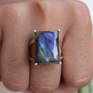 May include: A rectangular labradorite gemstone ring. The stone displays iridescent blue and purple hues. The ring has a silver band with small silver accents at each corner of the stone. The ring is shown on a hand.