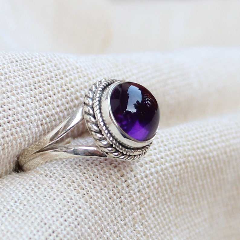 May include: A silver ring featuring a large, round, deep purple gemstone. The gemstone is set in a bezel with a twisted rope design around it. The ring's band splits into two at the top, adding a unique detail to the jewelry.