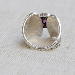 May include: A silver ring with a purple gemstone set in the center. The ring has a detailed design on the band.